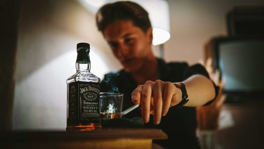 A moody image of a man smoking with a glass of whiskey indoors.