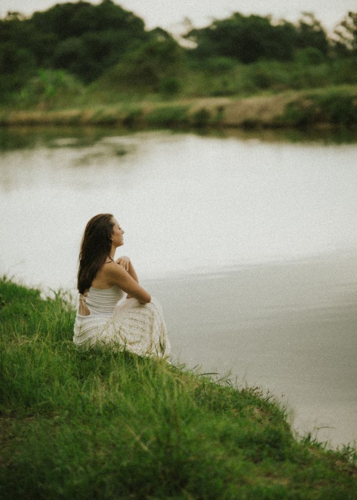 A peaceful moment captured as a woman in a white dress sits contemplatively by a calm lake.