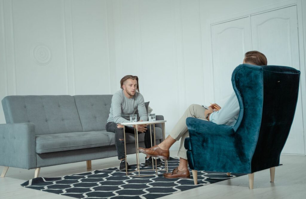 Two men engaged in a therapy session inside a modern office setting, discussing mental health.